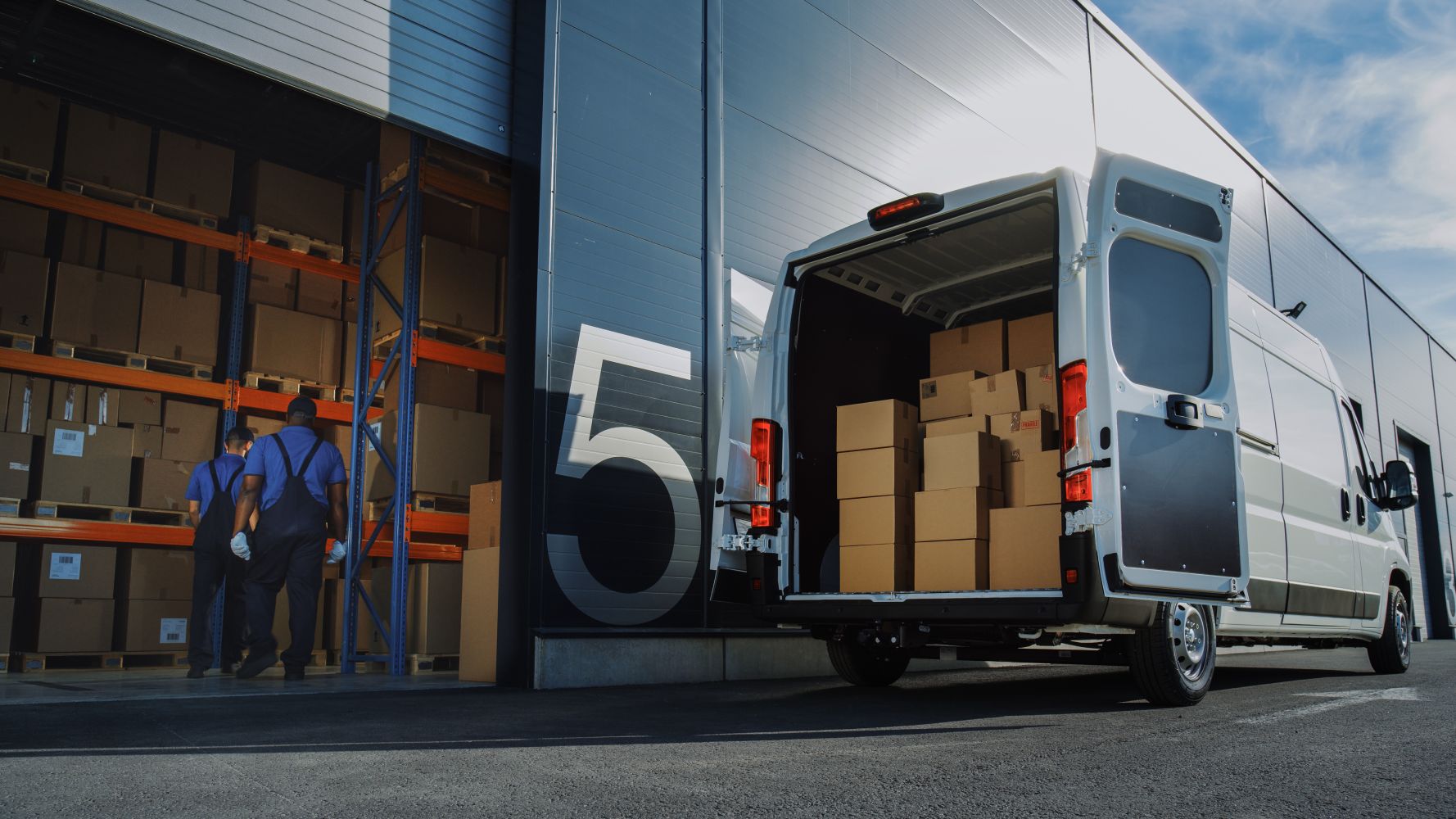 A delivery van loaded with boxes is parked outside a warehouse dock. Two workers in blue uniforms stand near the open door. The number "5" is visible.